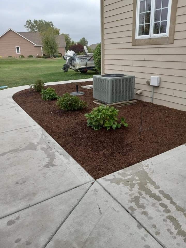 Un macizo de mantillo café alrededor de una unidad de aire acondicionado al costado de la casa, recién plantado con hortensias y rematado contra el concreto
