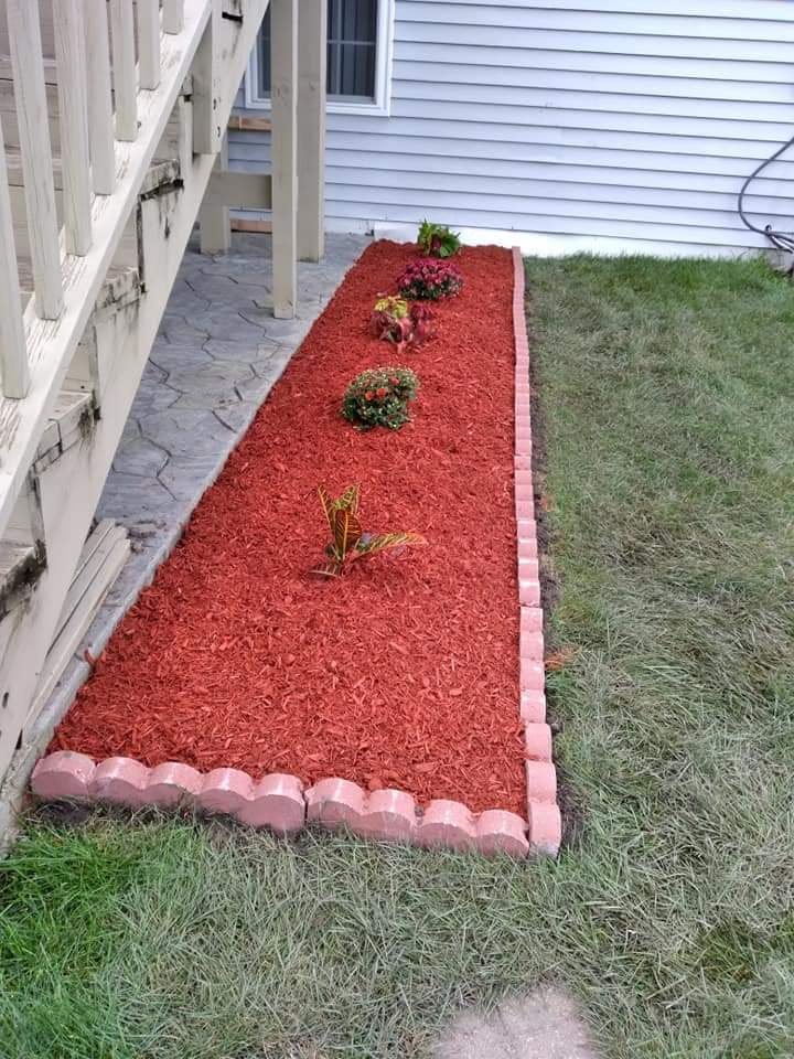Un macizo largo de mantillo rojo con borde de ladrillo ondulado junto a un patio de concreto estampado y las escaleras del deck
