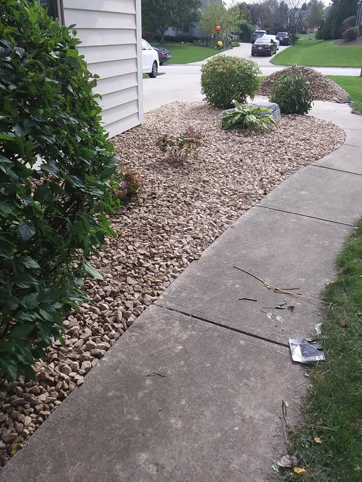 Un macizo de piedra decorativa beige a lo largo de la banqueta del frente y la entrada, plantado con arbustos recortados y hostas