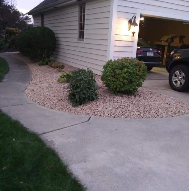 Un macizo curvo de piedra decorativa beige junto a un garaje y una entrada, plantado con arbustos recortados, iluminado por un farol al anochecer