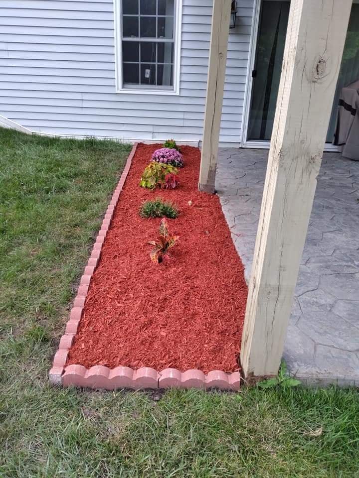 Un macizo nuevo de mantillo rojo bordeado con ladrillo ondulado junto a un patio de concreto estampado, plantado con crisantemos y coleos