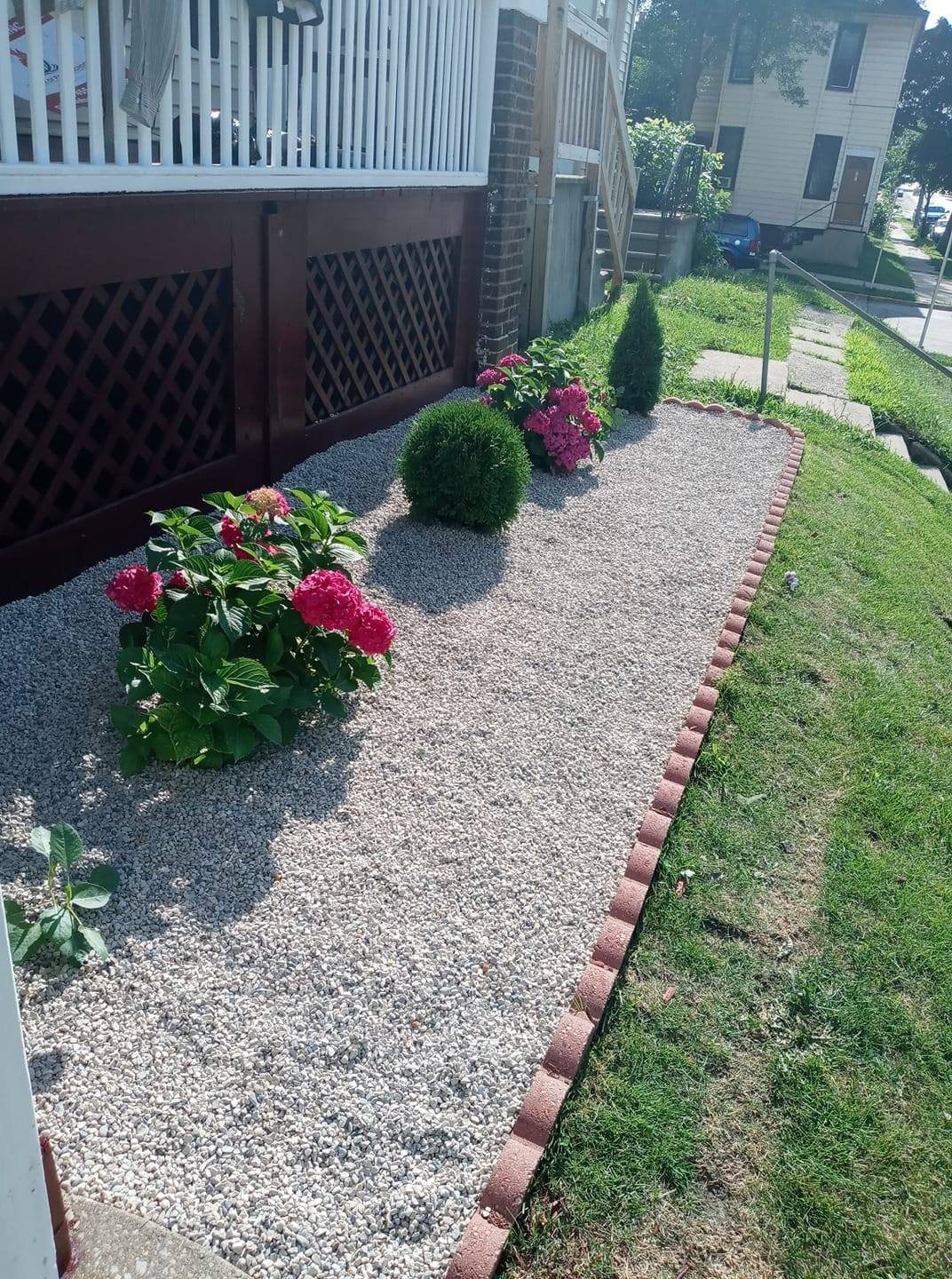 Un jardín lateral angosto cubierto con piedra decorativa gris y blanca, bordeado con ladrillo rojo ondulado y plantado con hortensias y una tuya en forma de globo
